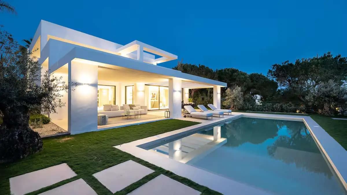 Modern white villa at dusk, with a lit terrace beside the pool